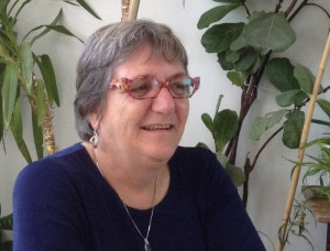 Photo de madame Provencher : lunettes stylisées un peu papillon couleur saumon ; cheveux semi-courts, souriante. Plusieurs plantes de maison derrière.